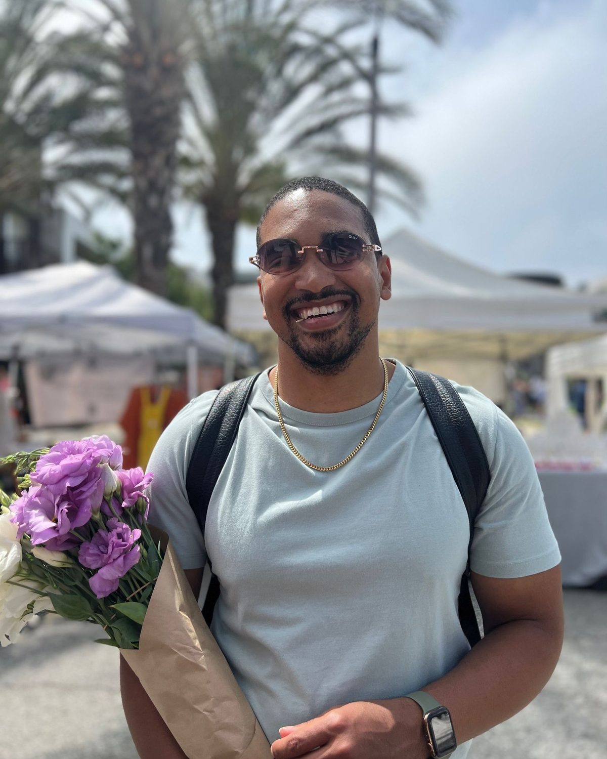 Jared laughing at a farmers market, sunglasses on, holding a bunch of purple flowers